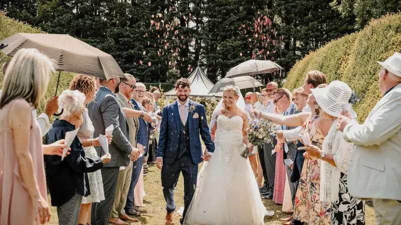 Bride and groom walking through a garden between guests holding umbrellas.